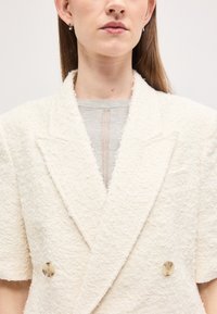 Femme portant un blazer à manches courtes texturé blanc cassé par-dessus un haut gris clair avec un détail de couture verticale, boucle d'oreille argentée visible.