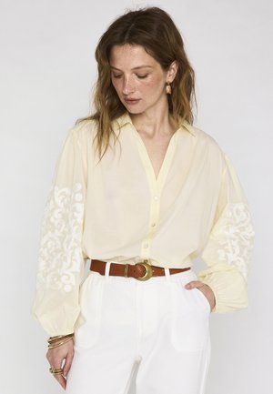 Vrouw draagt lichtgele blouse met geborduurde pofmouwen en witte broek, hand in de zak, kijkt naar beneden, accessoires met bruine riem en gouden sieraden.