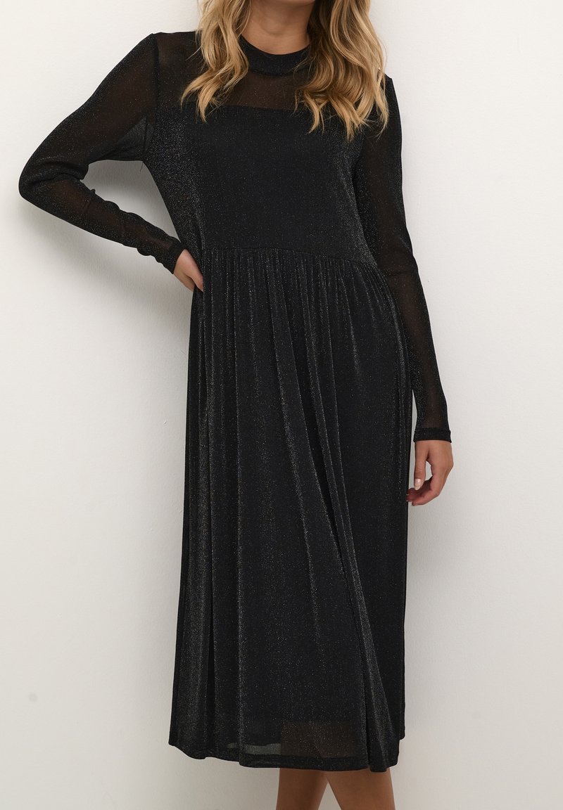 Femme portant une longue robe noire scintillante avec des manches longues transparentes, debout contre un mur blanc uni, main posée sur la hanche.