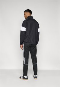 Μαύρο αθλητικό μπουφάν με λευκές λεπτομέρειες, σε συνδυασμό με μαύρο παντελόνι φόρμας με λευκές ρίγες και μαύρα αθλητικά παπούτσια με λευκό λογότυπο.