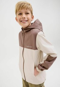Veste coupe-vent avec un design bicolore, le haut est marron et le bas est beige, équipée d'une capuche et de poignets élastiques.