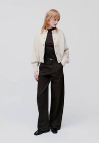 Cardigan de tricô cor de creme com botões, usado sobre um suéter de gola alta preto. Combinado com calças largas pretas e sapatos pretos.