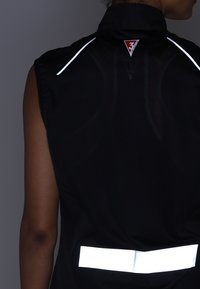 Czarna bezrękawnik z odblaskowymi białymi akcentami, z siateczkowymi wstawkami i logo na karku z tyłu dla lepszej widoczności.