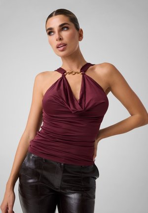 Donna con top smanicato bordeaux con dettaglio catena dorata e pantaloni in pelle nera testurizzata, che posa con una mano sul fianco.