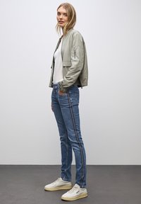 Kobieta stoi w casualowym stroju z jasnoszarym bomberem, białą koszulą, niebieskimi dżinsami z bocznymi paskami i białymi trampkami na szarym podłożu.