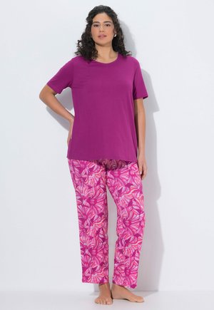 Kobieta stoi boso, ubrana w jednolity fioletowy krótki rękaw i luźne różowe spodnie w kwiatowy wzór na białym tle.