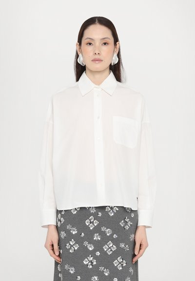 Chemise blanche à manches longues, avec un col pointu, une poche poitrine simple sur le côté gauche et une coupe décontractée. Portée avec une jupe grise ornée.