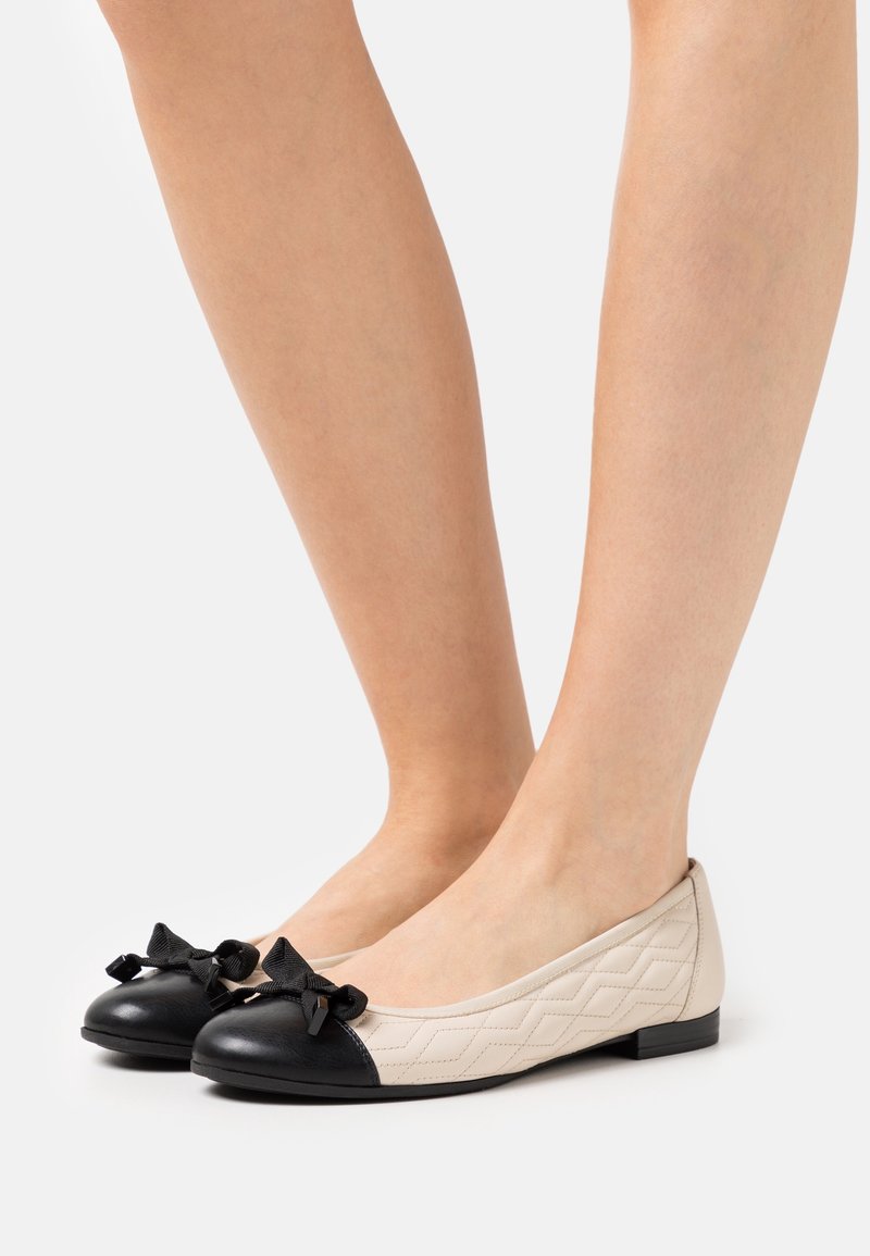 Jambes portant des ballerines matelassées crème avec embouts noirs et nœuds décoratifs noirs, se tenant sur une surface claire unie.