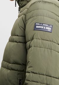 Zielona kurtka puchowa z pikowanym wzorem, z zasuwaną kieszenią i czarną metką z logo "SCOTCH & SODA" na ramieniu.