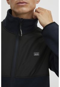 Man draagt een marineblauwe fleecejas met een zwart borstpaneel, stelt de kraagrits bij waarop de tekst "11 Project" en een logovlek op de borst te zien zijn.