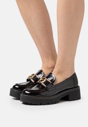 Mocassins en cuir verni noir brillant avec une semelle épaise noire, présentant une boucle en métal doré sur le dessus et un design de bout arrondi.