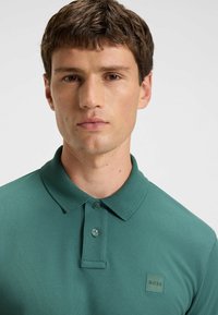 Polo in verde scuro, maniche corte, tessuto strutturato, collo con due bottoni e un piccolo logo quadrato sul lato sinistro del petto.