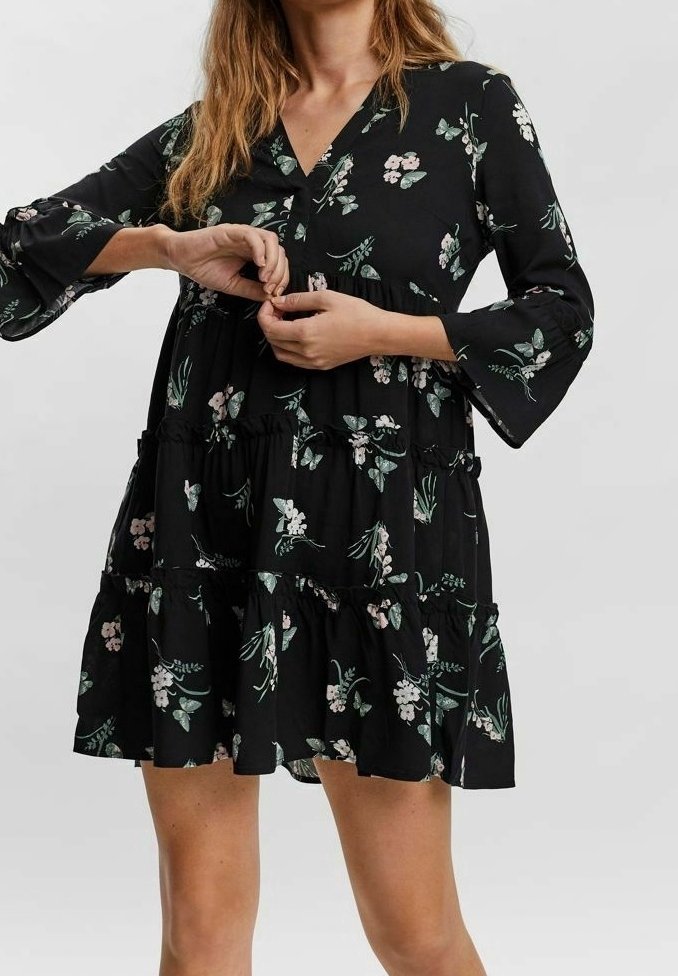Femme portant une robe noire fleurie à manches longues et jupe à volants, boutonnant les boutons au niveau de la poitrine.