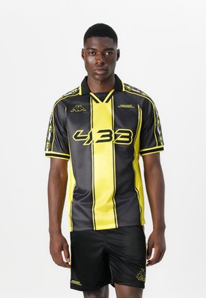 Črno- Rumena športna majica z kratkimi rokavi, ki vsebuje navpične črte, ovratnik in drzne grafike. Vključene so črne kratke hlače.