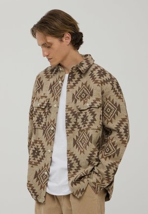 Camicia marrone a fantasia in una calda tonalità beige con disegni geometrici, due tasche sul petto e bottoni bianchi, indossata sopra una t-shirt bianca.