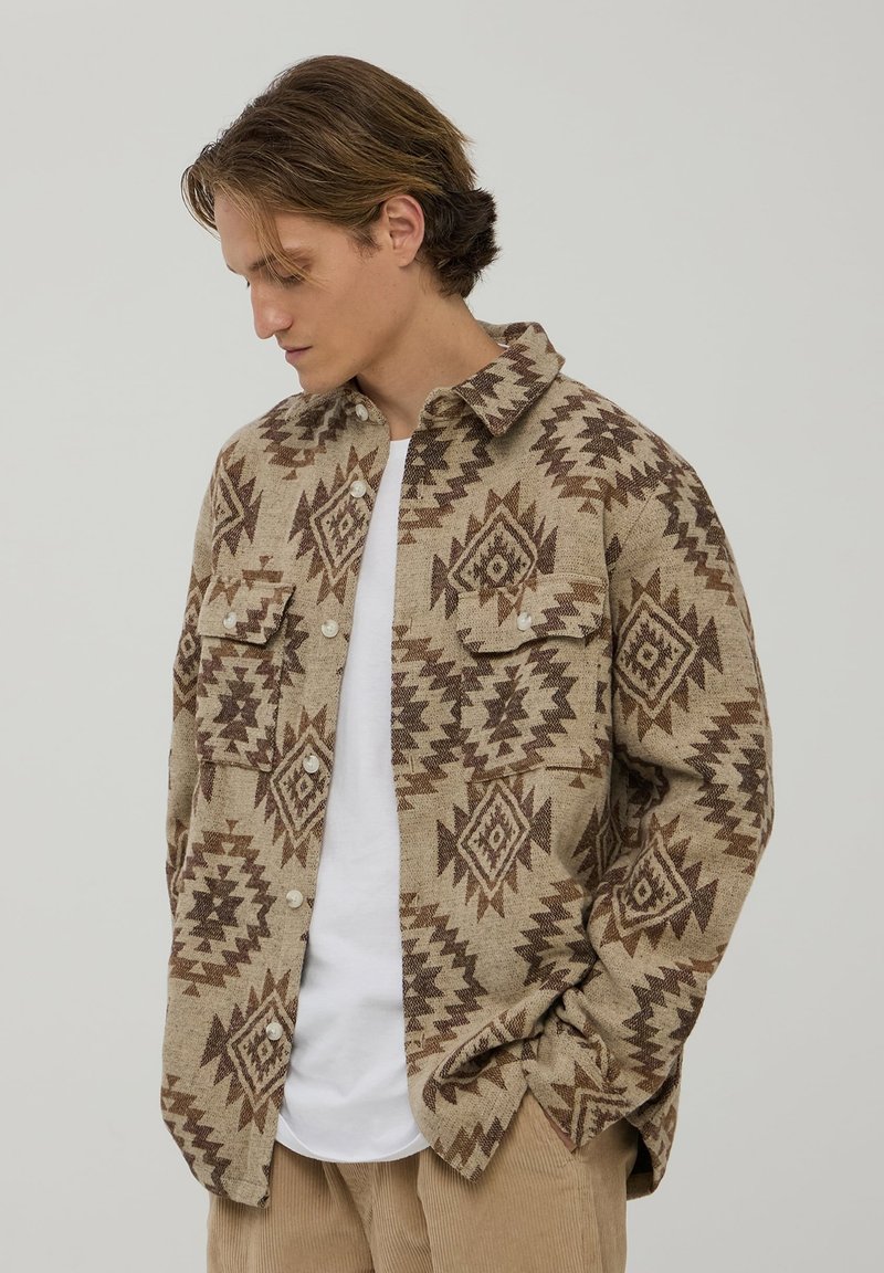 Brązowa koszula w beżowym odcieniu z geometrycznymi wzorami, z dwoma kieszeniami na piersiach i białymi guzikami, noszona na białej koszulce.