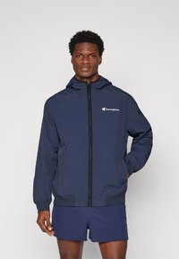 Navy hooded jacket made of lightweight material, featuring a front zipper and logo on the left chest. Smooth texture with elastic cuffs.