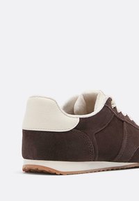 Sneaker in suede marrone a profilo basso con inserto beige sul tallone, collo imbottito, soletta bianca e suola in gomma colorata, mostrato da un angolo laterale posteriore.