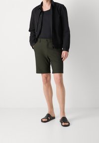 Černá lehká zipová bunda, padnoucí černé tílko, olivově zelené šortky a černé sandály. Hladké textury a ležérní design.