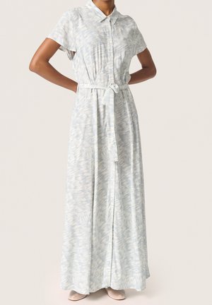 Mujer con vestido largo de manga corta, color gris claro con estampado, con cuello, abotonado en la parte delantera y cinturón atado a la cintura, de pie con las manos en las caderas.