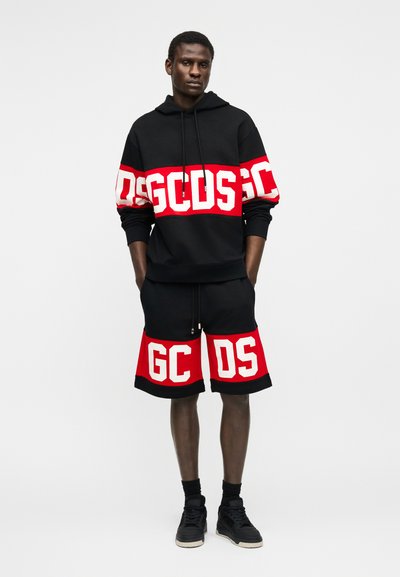 Uomo con felpa con cappuccio nera e pantaloncini con scritte audaci rosse e bianche "GCDS", in piedi con le mani nelle tasche su uno sfondo semplice.