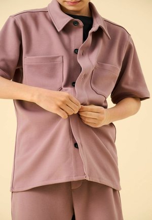 Žmogus sagsto dulkinės rožinės spalvos trumpomis rankovėmis marškinius su priekinėmis kišenėmis, dėvi derančias kelnes, ant smėlio spalvos fono.