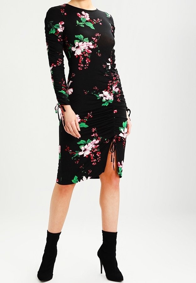 Femme portant une robe noire à manches longues, longueur genou, avec un imprimé floral rose et vert et des bottines noires à talons hauts.