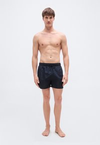 Μαύρο μαγιό βερμούδα με κορδόνι στη μέση, κατασκευασμένο από λείο υλικό. Το μοντέλο στέκεται ξυπόλητο, παρουσιάζοντας έναν μινιμαλιστικό σχεδιασμό.