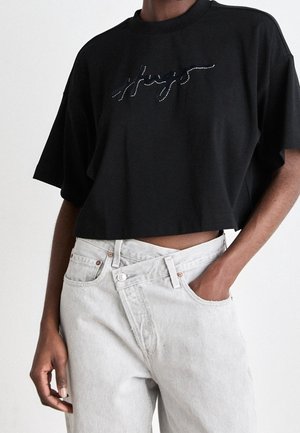 Persoon draagt een zwart cropped t-shirt met een gestikt ontwerp en lichtgrijze high-waisted jeans met een asymmetrische knoopsluiting.