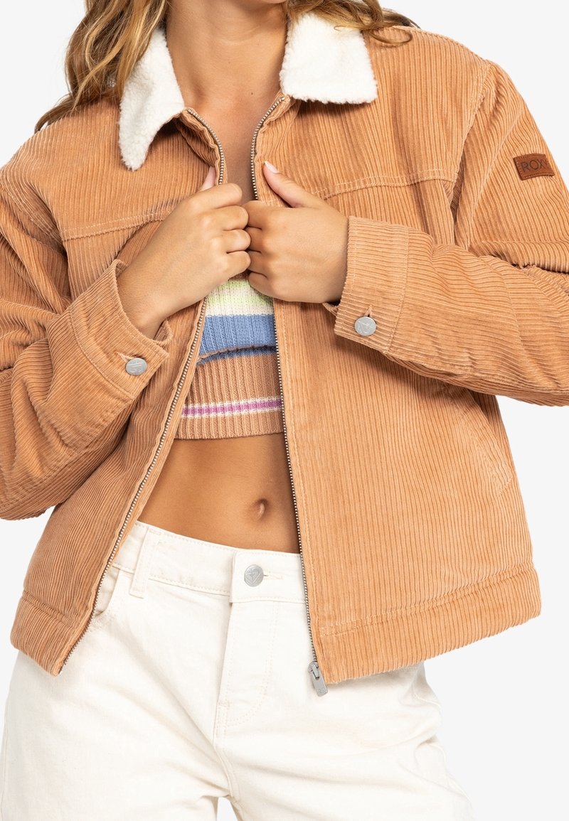 Veste en velours côtelé couleur fauve avec un col en fausse fourrure blanche. Présente une fermeture éclair à l'avant, une texture côtelée et des pressions métalliques. Portée sur un haut en tricot coloré.