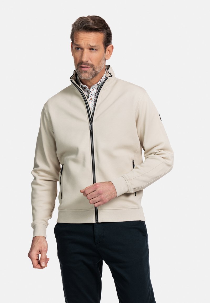 Beige ritsjack met een hoge kraag, voorzien van zwarte ritsaccenten en zijzakken, gedragen over een geblokt shirt. Zachte stoftextuur.