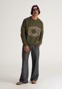 Olive green knitted sweater with a beige circular pattern, paired with black and white striped wide-leg pants and beige moccasin shoes.