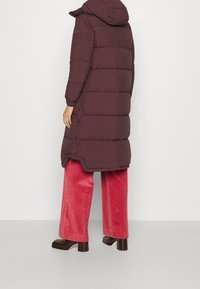 Bordeauxrote Pufferjacke mit Kapuze, gestepptem Design und Druckknopfverschluss, kombiniert mit Schlagjeans aus rotem Cord und schwarzen klobigen Stiefeletten.
