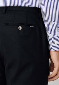 Crne krojene hlače s teksturiranim završetkom, sa stražnjim džepom s gumbom i etiketom na kojoj piše "HACKETT." Vidljiva prugasta košulja.