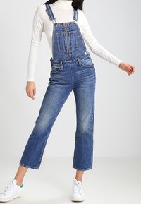 Denim overalls with a straight leg cut, adjustable straps, and front pockets. Worn over a white turtleneck and complemented by white sneakers.
