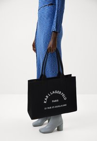Borsa tote in tela nera con doppie maniglie, caratterizzata da un testo bianco in grassetto e una forma strutturata; portata insieme a un vestito blu a coste e stivali grigi.