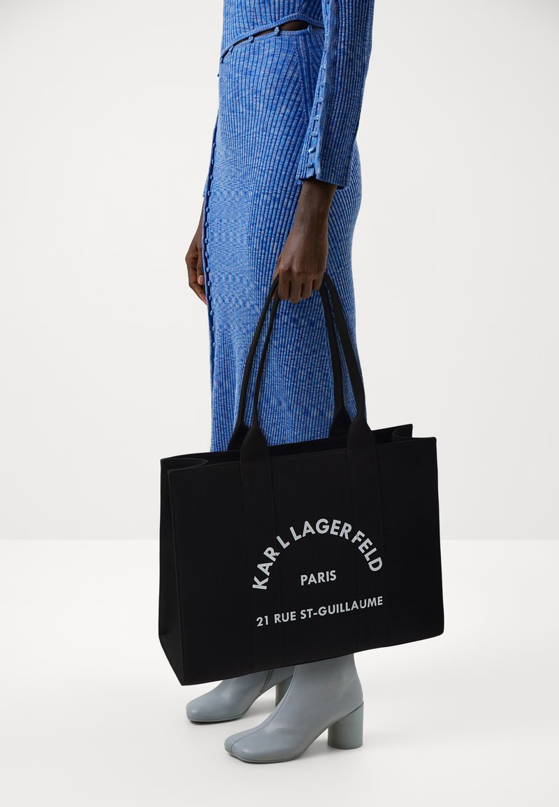 Borsa tote in tela nera con doppie maniglie, caratterizzata da un testo bianco in grassetto e una forma strutturata; portata insieme a un vestito blu a coste e stivali grigi.