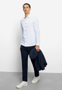 Weißes Hemd mit schwarzen Knöpfen, kombiniert mit marineblauer Hose. Modell hält eine marineblaue Jacke und trägt weiße Turnschuhe. Glatter Stoff, maßgeschneiderte Passform.