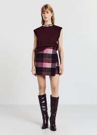 Top in maglia color bordeaux con scollo impreziosito da gioielli, abbinato a una gonna a quadri rosa e nera. Stivali bordeaux lucidi fino al ginocchio completano il look.