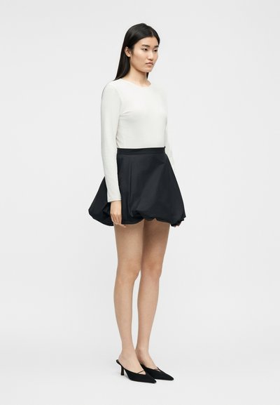 Femme debout portant un haut blanc à manches longues, une mini-jupe noire bouffante et des escarpins noirs à talons petits et pointus, sur un fond uni.