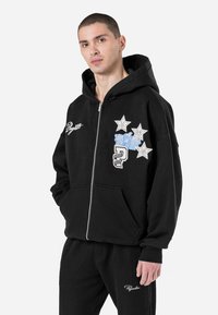 Crna zip-up hoodie s velikom kapuljačom, prednjim džepovima i izvezenim dizajnom u plavoj i bijeloj boji. Izrađena od mekane, teksturirane tkanine.
