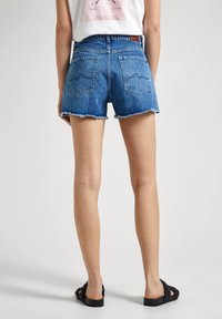 Denim šortky s roztrhnutým okrajom, modrým prešitím a dvoma zadnými vreckami s jemným vlnitým vzorom. Kombinované s čiernymi sandálmi.