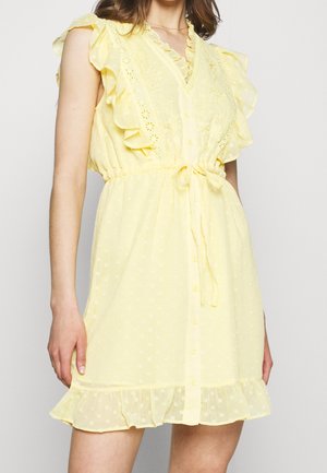 Kobieta ubrana w żółtą sukienkę bez rękawów z falbanami, haftowanymi kwiatami, wiązaniem w talii i zapinana na guziki z przodu.