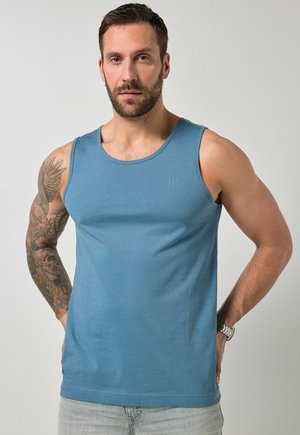 Man with beard and tattooed left arm wearing a blue sleeveless shirt and light jeans, hands resting at his hips against a plain background.