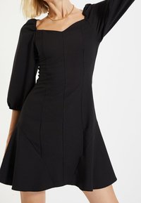 Vestido negro con escote cuadrado, corsé ajustado y mangas largas abullonadas. Presenta costuras verticales y una falda acampanada.