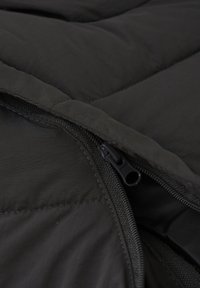 Giacca a vento nera in tessuto imbottito con design trapuntato. Presenta una cerniera nera e una texture liscia, con cuciture visibili lungo i bordi.