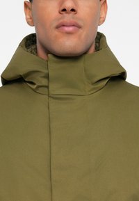 Mann, der eine olivgrüne Jacke mit hohem Kragen trägt, zeigt nur das untere Gesicht und den Hals vor einem einfarbigen Hintergrund.