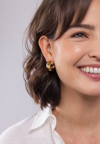 Goldene Kreolenohrringe mit strukturierter Oberfläche, prominent an einem Modell mit kurzen, lockigen Haaren zu sehen, das ein weißes Hemd mit Kragen trägt.