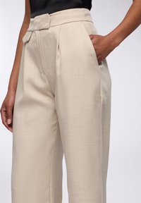 Pantaloni beige su misura con finitura testurizzata, dotati di vita alta, tasche laterali e un design frontale piatto.