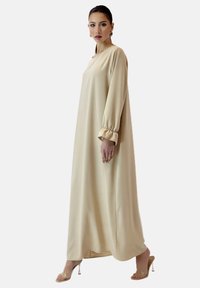 Beige langes Kleid mit lockerer Passform, gerafften Bündchen und glatter Textur. Der Saum fällt elegant und wird mit klaren Absatzsandalen kombiniert.